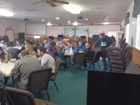 People seated at tables in a community hall, engaging in conversation and activities. The room has a mural, rectangular tables, chairs, and ceiling fans. {{brizy_dc_image_alt imageSrc=